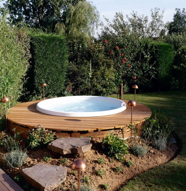 Whirlpool Gartengestaltung mit rundem Außenwhirlpool auf einer erhöhten Holzterrasse, umgeben von Hecken, Bäumen und Gartenbeleuchtung