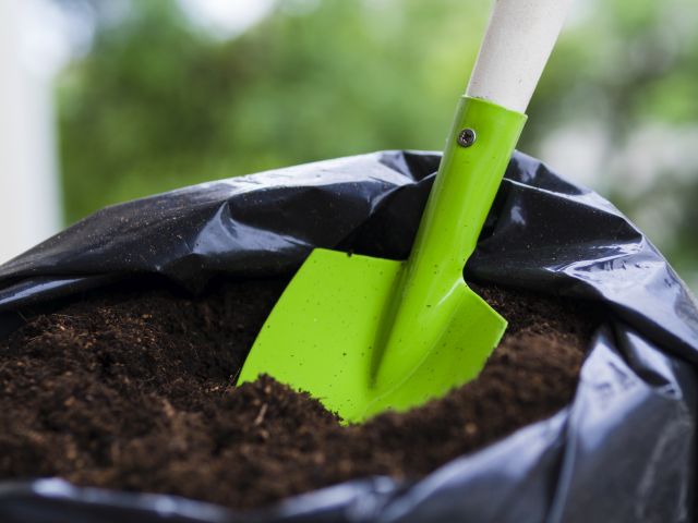 Grüne Gartenschaufel steckt in dunkler Blumenerde in einem mit schwarzer Gartenfolie ausgekleideten Pflanzbehälter – Vorbereitung von Erde für Pflanzen im Garten oder Gewächshaus