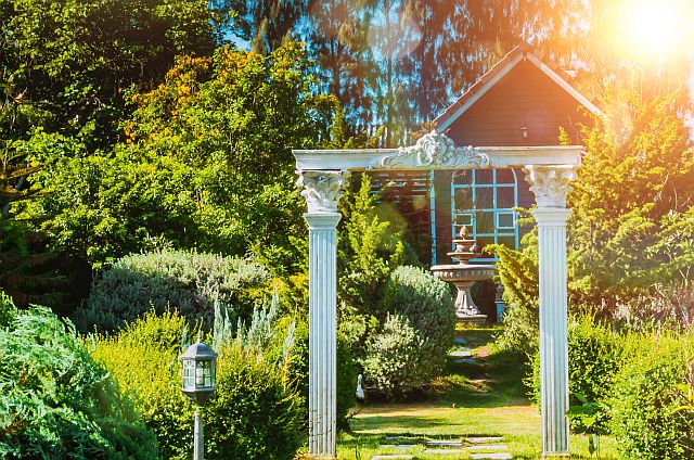 Klassische weiße Pergola mit dekorativen Säulen als Gartentor in einer gepflegten Gartenanlage, umgeben von üppigem Grün, Sträuchern und Bäumen, romantische Gartenarchitektur mit Sonnenlicht im Hintergrund