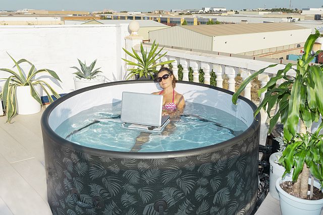 Moderner Outdoor Whirlpool auf Dachterrasse, genutzt als Homeoffice im Wasser – Person sitzt im warmen Whirlpool mit Laptop, umgeben von Pflanzen, ideal für Entspannung, Wellness und Arbeiten im Freien