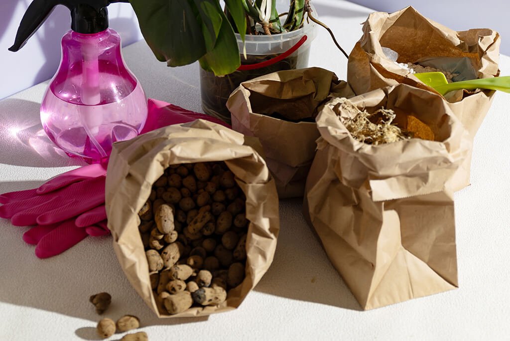 Große braune Samentüten aus Papier mit verschiedenen Pflanzsubstraten wie Tongranulat und Erde, daneben Gartenhandschuhe, eine rosa Sprühflasche und ein Blumentopf mit Orchidee auf hellem Tisch
