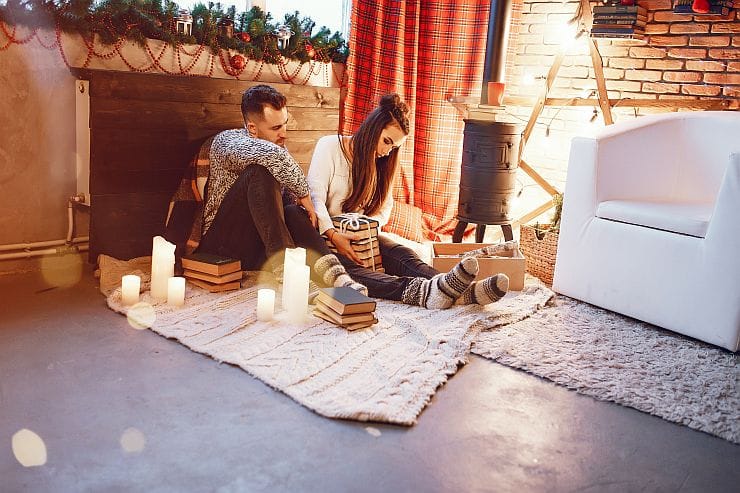 Paar sitzt auf einer Terrasse auf Decken neben Kerzen und Holzofen und wärmt sich an einem gemütlichen Winterabend